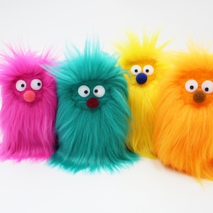 May include: Four colorful, fuzzy monster plush toys. The plush toys are pink, teal, yellow, and orange. Each has large googly eyes, a small nose, and a shaggy, furry body. The toys are standing upright against a white background.
