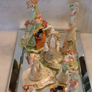 K&ouml;nnte beinhalten: Eine Sammlung skurriler Keramikfiguren, darunter ein Zauberer, Schneem&auml;nner und Blumenskulpturen, die auf einer verspiegelten Oberfl&auml;che ausgestellt sind. Die Figuren weisen Pastellfarben, florale Akzente und komplizierte Details auf. Einige Figuren haben Etiketten mit der Aufschrift "Stinging Bee Pottery".