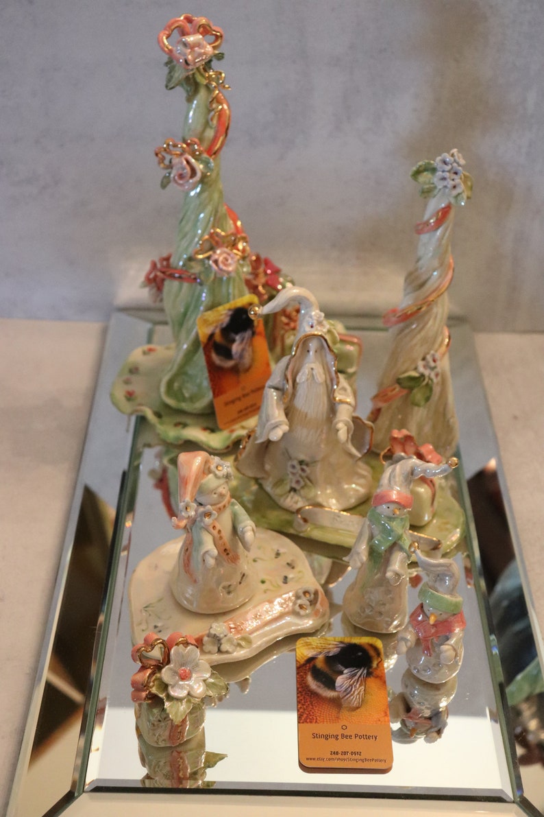 K&ouml;nnte beinhalten: Eine Sammlung skurriler Keramikfiguren, darunter ein Zauberer, Schneem&auml;nner und Blumenskulpturen, die auf einer verspiegelten Oberfl&auml;che ausgestellt sind. Die Figuren weisen Pastellfarben, florale Akzente und komplizierte Details auf. Einige Figuren haben Etiketten mit der Aufschrift "Stinging Bee Pottery".