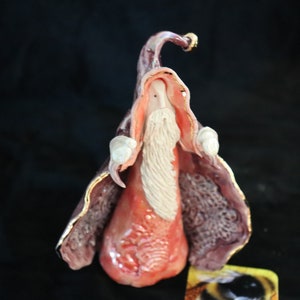 K&ouml;nnte beinhalten: Keramik-Zaubererfigur mit langem wei&szlig;em Bart, rotem Gewand und lila Umhang mit goldenen Verzierungen. Der Zaubererhut ist ebenfalls lila mit goldenen Akzenten. Die Figur ist vor einem dunklen Hintergrund platziert.