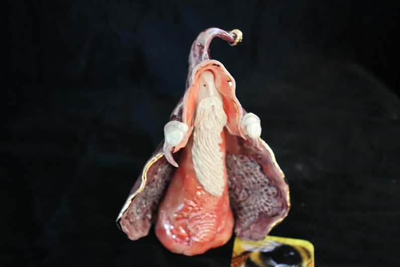 K&ouml;nnte beinhalten: Keramik-Zaubererfigur mit langem wei&szlig;em Bart, rotem Gewand und lila Umhang mit goldenen Verzierungen. Der Zaubererhut ist ebenfalls lila mit goldenen Akzenten. Die Figur ist vor einem dunklen Hintergrund platziert.