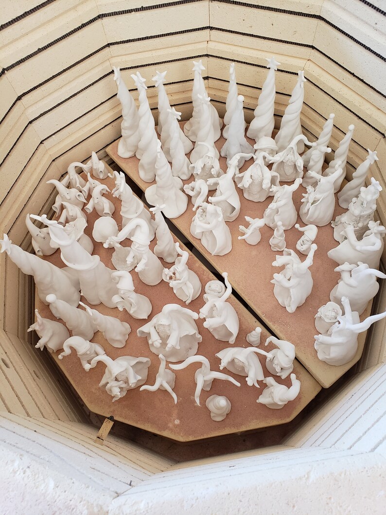 K&ouml;nnte beinhalten: Eine Sammlung wei&szlig;er Keramikfiguren, darunter Weihnachtsb&auml;ume und skurrile, zwergenartige Figuren, die in einem Ofen angeordnet sind. Die Figuren sind in verschiedenen Posen und Gr&ouml;&szlig;en, bereit zum Brennen.