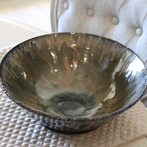 May include: A decorative ceramic bowl with a metallic glaze in shades of gray, brown, and green. The rim features a textured, rope-like design. The bowl is set on a textured surface, with a tufted chair in the background.