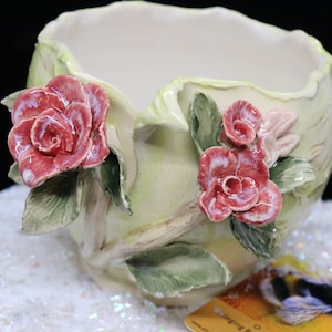 May include: A ceramic planter with a light green base and detailed red rose accents. The planter has a rounded shape with a scalloped rim and is decorated with sculpted roses and green leaves. The overall design is floral and decorative.