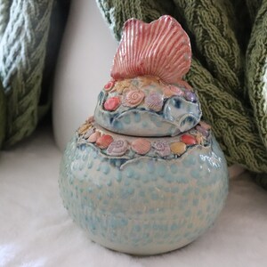 May include: A ceramic jar with a blue and white glaze and a lid decorated with seashells and a pink seashell finial.