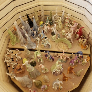 K&ouml;nnte beinhalten: Eine Sammlung von Keramikfiguren, darunter skurrile Zauberer- und Elfenfiguren sowie spiralf&ouml;rmige B&auml;ume, in einem Ofen. Die Figuren sind in verschiedenen Farben, darunter Gr&uuml;n, Blau und Wei&szlig;, auf einem Holzregal angeordnet.