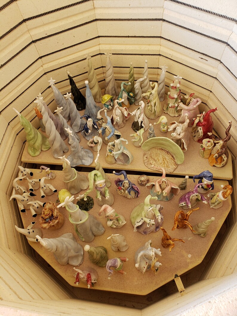 K&ouml;nnte beinhalten: Eine Sammlung von Keramikfiguren, darunter skurrile Zauberer- und Elfenfiguren sowie spiralf&ouml;rmige B&auml;ume, in einem Ofen. Die Figuren sind in verschiedenen Farben, darunter Gr&uuml;n, Blau und Wei&szlig;, auf einem Holzregal angeordnet.