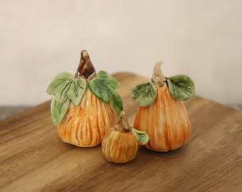 May include: Three ceramic pumpkins in various sizes, with orange and green painted details. The pumpkins are sitting on a wooden surface.