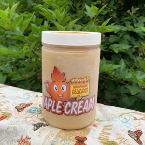 May include: A clear glass jar with a white lid filled with a light brown maple cream. The jar has a cartoon maple leaf with a smiling face and the text "Grandma's Homemade Delicious Maple Cream".