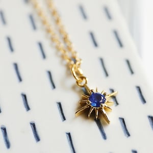 May include: A gold necklace with a blue gemstone star pendant. The necklace is on a white background with blue lines.