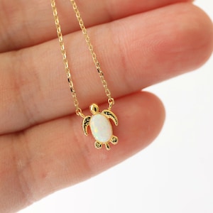 Può includere: Una delicata collana d'oro con un piccolo ciondolo a forma di tartaruga. Il ciondolo ha un centro simile all'opale e pinne e guscio dettagliati. La catena sottile è dorata. La collana è esposta su uno sfondo chiaro.