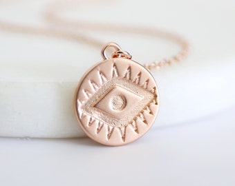 May include: A rose gold necklace with a round pendant featuring an evil eye design. The pendant has a raised, diamond-shaped eye with a smaller circle in the centre. The eye is surrounded by a circle of points.