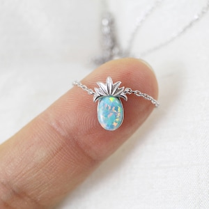 Op de afbeelding: Een zilveren ketting met een kleine hanger in de vorm van een ananas. De hanger heeft een bovenkant van gepolijst zilver en een basis van een ovale, iriserende blauwe opaalsteen. De ketting bevat een fijne zilveren ketting die aan de zijkanten is bevestigd.