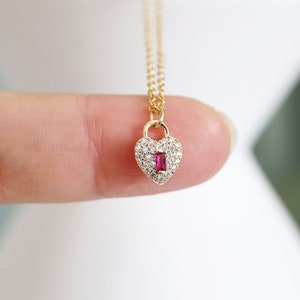 May include: A gold necklace with a heart-shaped pendant. The pendant is encrusted with small clear stones and has a single red stone in the center.