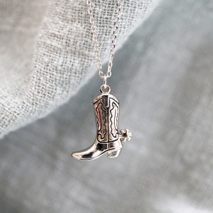 Könnte beinhalten: Eine silberfarbene Halskette mit einem Anhänger in Form eines Cowboy-Stiefels. Der Stiefel hat detaillierte, geätzte Muster und einen kleinen Sporn. Die zarte Kette hält den Anhänger vor einem weichen, hellblauen Hintergrund. Ein Schmuckstück im Western-Stil.