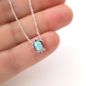Op de afbeelding: Een zilveren ketting met een schildpadhanger. De hanger heeft een lichtblauwe opaal midden en gedetailleerde vinnen en kop. De ketting is delicaat. De ketting wordt getoond tegen een vage achtergrond.