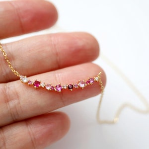 May include: A gold chain necklace with a delicate row of pink, red, and white gemstones. The gemstones are arranged in a crescent shape.