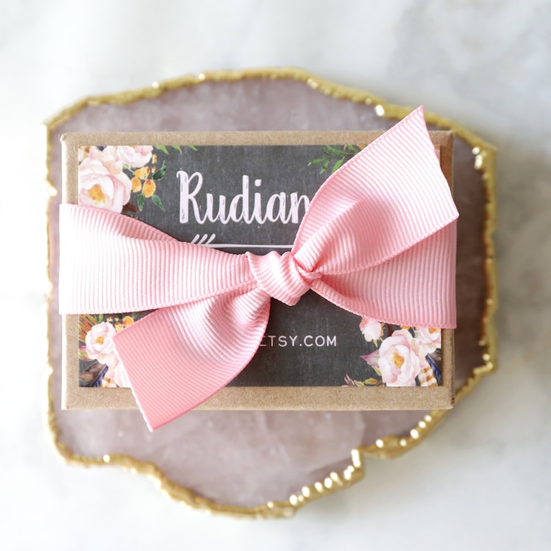 Op de afbeelding: Een klein, bruin cadeaudoosje met een roze lint dat in een strik is gebonden. Het doosje heeft een bloemenmotief en de tekst "Rudians Etsy.com" erop gedrukt.