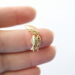 May include: A pair of gold-coloured fish earrings. Each earring features a small, detailed fish pendant hanging from a delicate gold wire. The fish have textured scales and fins. The earrings are displayed against a blurred white background.