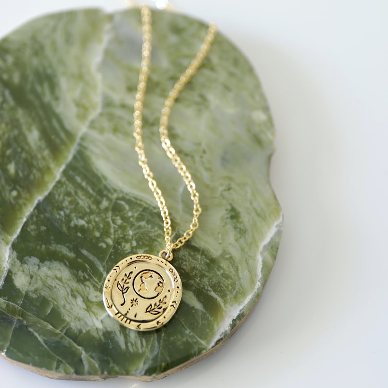 Op de afbeelding: Een gouden ketting met een ronde hanger met een gestileerd beeld van de aarde met bladeren en sterren. De hanger ligt op een groene marmeren ondergrond.