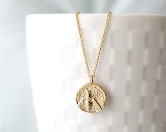 Peut inclure: Collier doré avec un pendentif rond représentant une abeille détaillée. Le pendentif est suspendu à une chaîne délicate. Le collier est présenté sur un fond blanc.