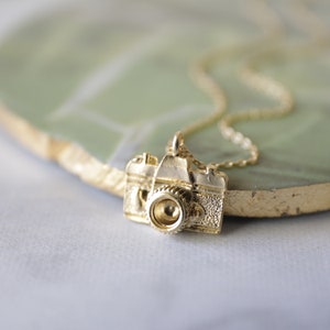 May include: A gold-colored necklace featuring a detailed pendant shaped like a vintage camera. The camera pendant has intricate detailing, including a lens and textured body. The necklace chain is delicate and also gold-colored.