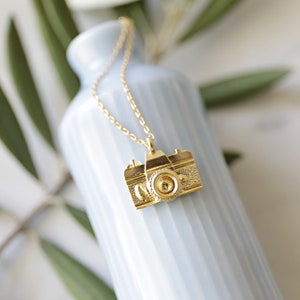 May include: A gold-colored necklace with a camera pendant. The pendant is a detailed miniature of a vintage camera, with textured surfaces and a prominent lens. The necklace hangs against a light blue vase, with green leaves in the background.