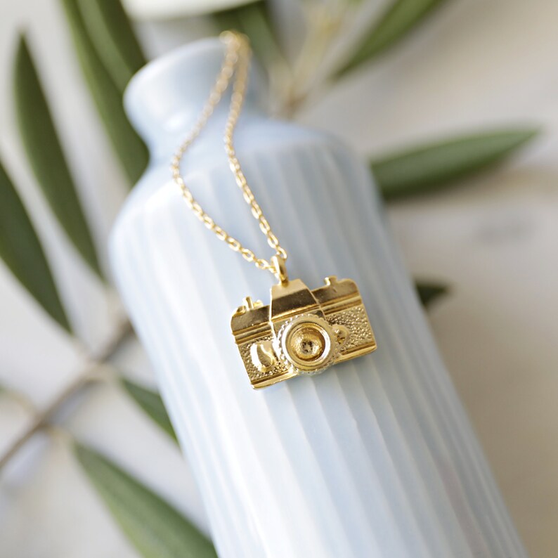 May include: A gold-colored necklace with a camera pendant. The pendant is a detailed miniature of a vintage camera, with textured surfaces and a prominent lens. The necklace hangs against a light blue vase, with green leaves in the background.