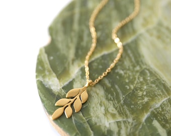 Könnte beinhalten: Eine goldfarbene Halskette mit einem zarten Anhänger in Form eines kleinen Zweigs mit mehreren glatten, abgerundeten Blättern. Die Kette ist dünn und metallisch und mit dem blattförmigen Anhänger verbunden, der ein mattes Finish aufweist.