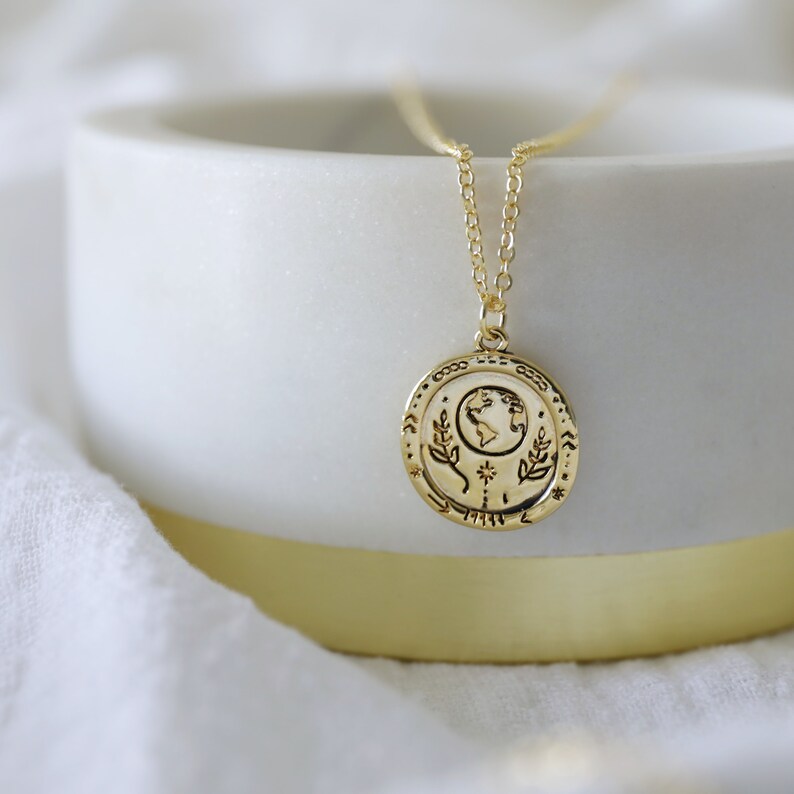Op de afbeelding: Een gouden ketting met een ronde hanger met een gedetailleerd beeld van de aarde, omringd door bladeren en een ster. De hanger ligt op een wit oppervlak.