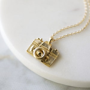 May include: A gold-colored camera pendant necklace. The pendant is a detailed miniature of a vintage camera, with a lens, buttons, and textured body. The necklace chain is delicate and gold, complementing the pendant's design.