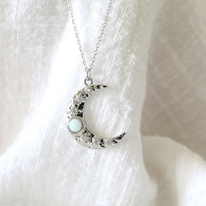 May include: Silver crescent moon pendant necklace featuring a white gemstone. The moon has a textured surface with small, sparkling details. The necklace hangs from a fine chain, a celestial-themed accessory.