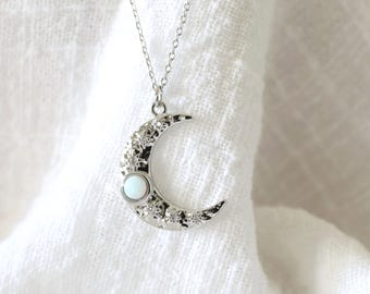 Könnte beinhalten: Silberfarbene Halskette mit einem Mond-Anhänger in Form einer Mondsichel und einem weißen Edelstein in der Mitte. Der Mond ist strukturiert mit kleinen, funkelnden Akzenten. Die Halskette hängt an einer zarten Kette.