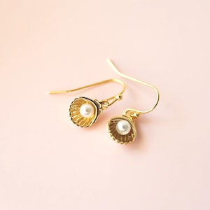 May include: A pair of gold-colored seashell earrings with a small white pearl in the center of each shell. The earrings have a hook closure and are set against a soft pink background.