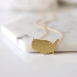 May include: A gold-colored necklace featuring a pendant in the shape of the United States. The pendant has a textured surface and is suspended from a delicate gold chain. The necklace is displayed on a white surface.