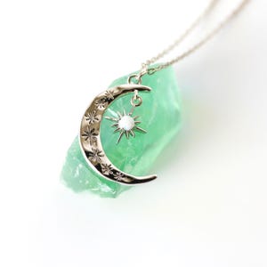 May include: A silver-toned crescent moon pendant necklace featuring engraved star motifs and a dangling star charm with a central white opal-like stone. The pendant rests against a rough, translucent light green mineral specimen.