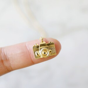 May include: A small, gold-colored camera pendant. The pendant is a detailed miniature replica of a classic camera, with a lens, viewfinder, and textured body. The camera is suspended from a delicate chain, suggesting it's a necklace.