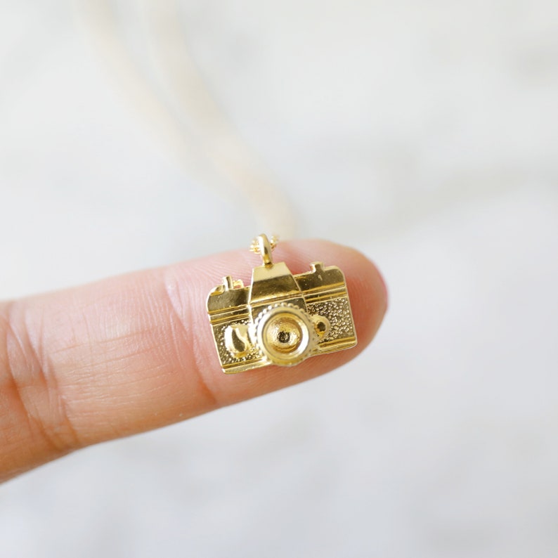 May include: A small, gold-colored camera pendant. The pendant is a detailed miniature replica of a classic camera, with a lens, viewfinder, and textured body. The camera is suspended from a delicate chain, suggesting it's a necklace.