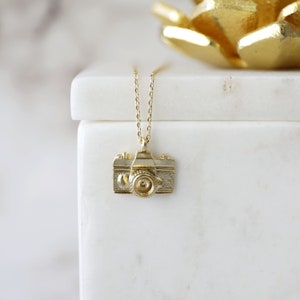 May include: A gold-colored camera pendant necklace. The pendant is a detailed miniature replica of a vintage camera. The necklace hangs on a delicate chain. The camera pendant is displayed on a white surface.