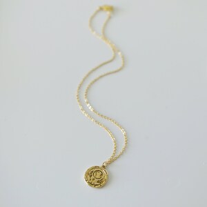 Op de afbeelding: Een gouden ketting met een ronde hanger met een maan en een bloemenmotief. De hanger heeft een inscriptie rond de rand.