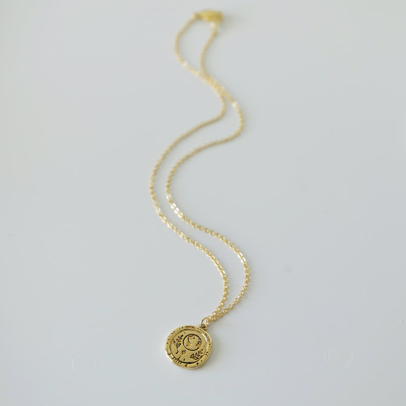 Op de afbeelding: Een gouden ketting met een ronde hanger met een maan en een bloemenmotief. De hanger heeft een inscriptie rond de rand.