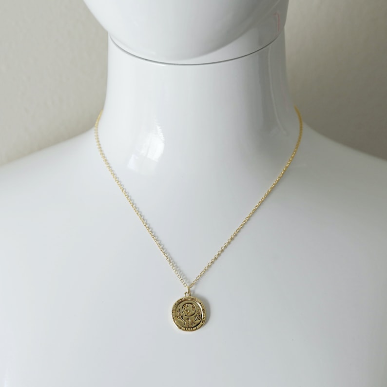 Op de afbeelding: Een gouden ketting met een ronde hanger met een gedetailleerd ontwerp van een halve maan en sterren. De hanger heeft een inscriptie rond de rand.