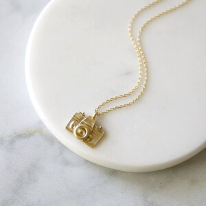 May include: A gold-colored necklace featuring a pendant shaped like a vintage camera. The camera pendant has detailed features, including a lens and textured body. The necklace chain is delicate and also gold-colored. The necklace is displayed on a white marble surface.