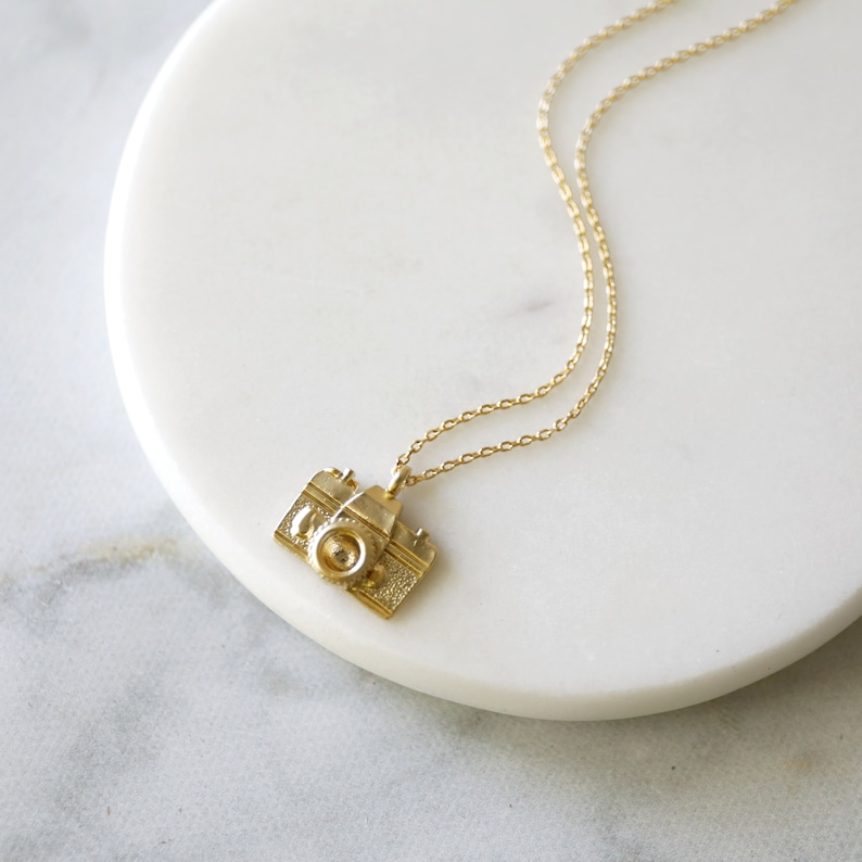 May include: A gold-colored necklace featuring a pendant shaped like a vintage camera. The camera pendant has detailed features, including a lens and textured body. The necklace chain is delicate and also gold-colored. The necklace is displayed on a white marble surface.