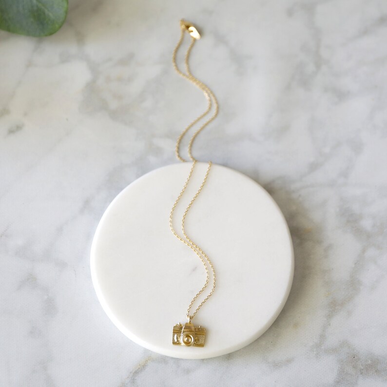 May include: A gold-colored necklace with a camera pendant. The necklace is displayed on a white, circular surface against a marble background. The camera pendant has detailed features, including a lens and buttons.