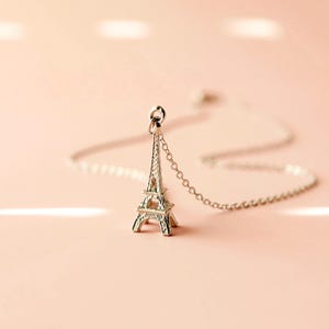 Puede incluir: Un collar de plata con un pequeño dije de la Torre Eiffel. El collar está sobre un fondo rosa.