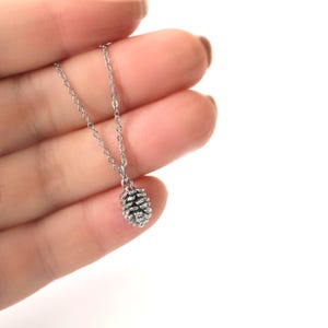 Könnte beinhalten: Eine silberfarbene Halskette mit einem kleinen Tannenzapfen-Anhänger. Der Anhänger ist detailliert und zeigt die Textur des Zapfens. Die Kette ist filigran und aus einem ähnlichen silberfarbenen Metall gefertigt. Die Halskette wird vor weißem Hintergrund gehalten.