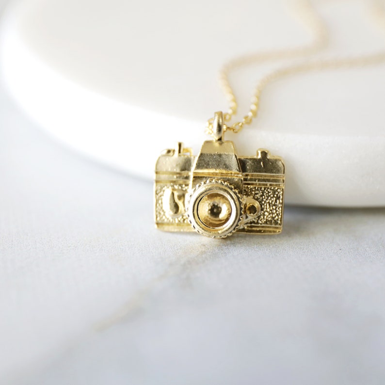 May include: A gold-colored camera pendant necklace. The pendant is a detailed miniature of a vintage camera, with textured surfaces and a prominent lens. The necklace chain is also gold, creating a cohesive and stylish accessory.