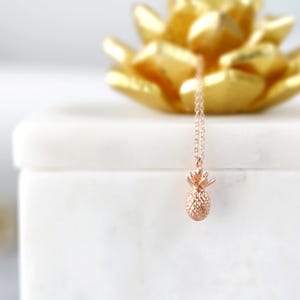 May include: A rose gold pineapple pendant necklace on a white marble surface. The necklace is delicate and features a small, detailed pineapple charm.