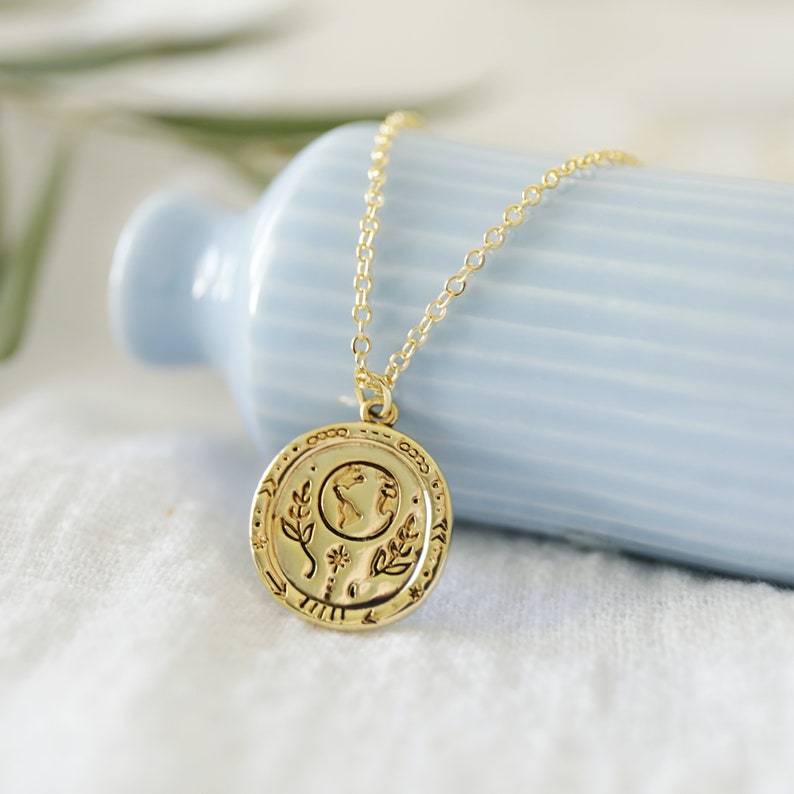 Op de afbeelding: Een gouden ketting met een ronde hanger met een gedetailleerd ontwerp van de aarde, bladeren en een bloem. De hanger zit aan een delicate ketting.